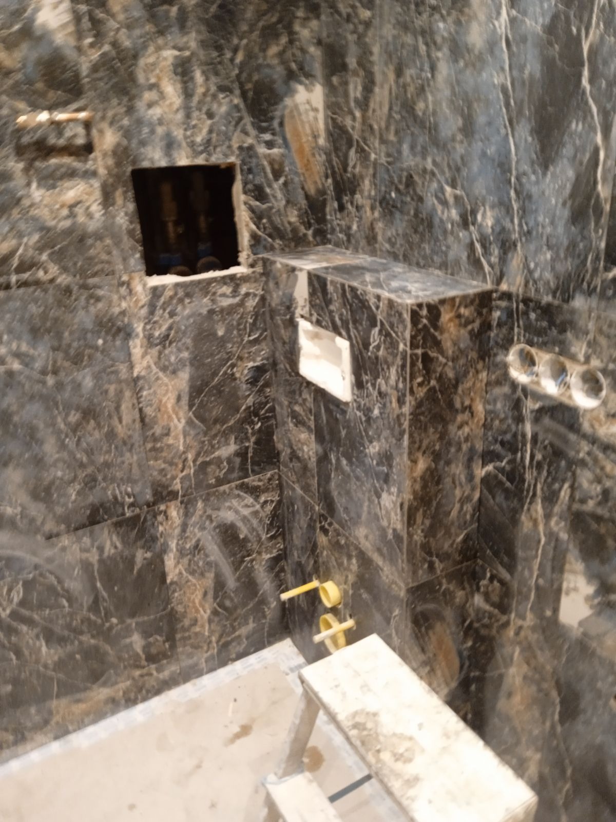 Łazienka w trakcie wykańczania z płytkami imitującymi czarny marmur, widoczne rury, otwór na instalację oraz drabinka malarska.