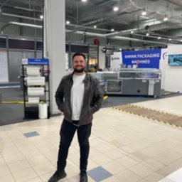 Uśmiechnięty mężczyzna w skórzanej kurtce stojący na tle hali z maszynami do pakowania folią termokurczliwą i regałem z folią, prawdopodobnie na targach lub wystawie.