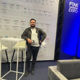 Uśmiechnięty mężczyzna w skórzanej kurtce stoi na tle stoisk targowych podczas PTAK Warsaw Expo, opierając się o biały stolik z telefonem w dłoni.