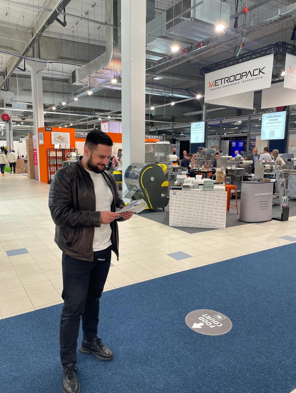 Mężczyzna w skórzanej kurtce przegląda ulotkę na targach maszyn pakujących Ptak Expo w Nadarzynie, na tle stoiska z maszynami do pakowania żywności.