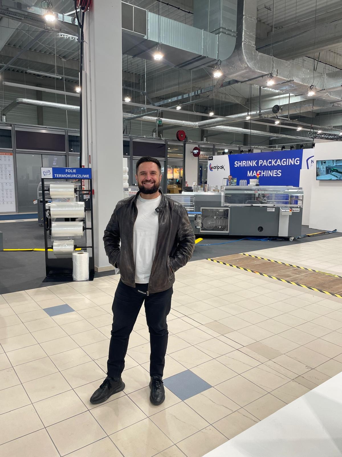 Uśmiechnięty mężczyzna w skórzanej kurtce stojący na tle hali z maszynami do pakowania folią termokurczliwą i regałem z folią, prawdopodobnie na targach lub wystawie.