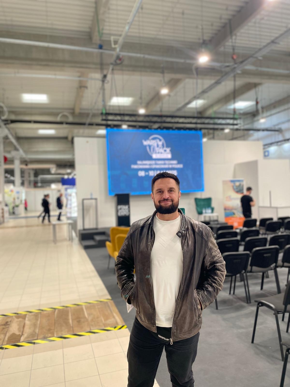 Mężczyzna w skórzanej kurtce na tle sceny targowej z ekranem wyświetlającym logo i nazwę wydarzenia.