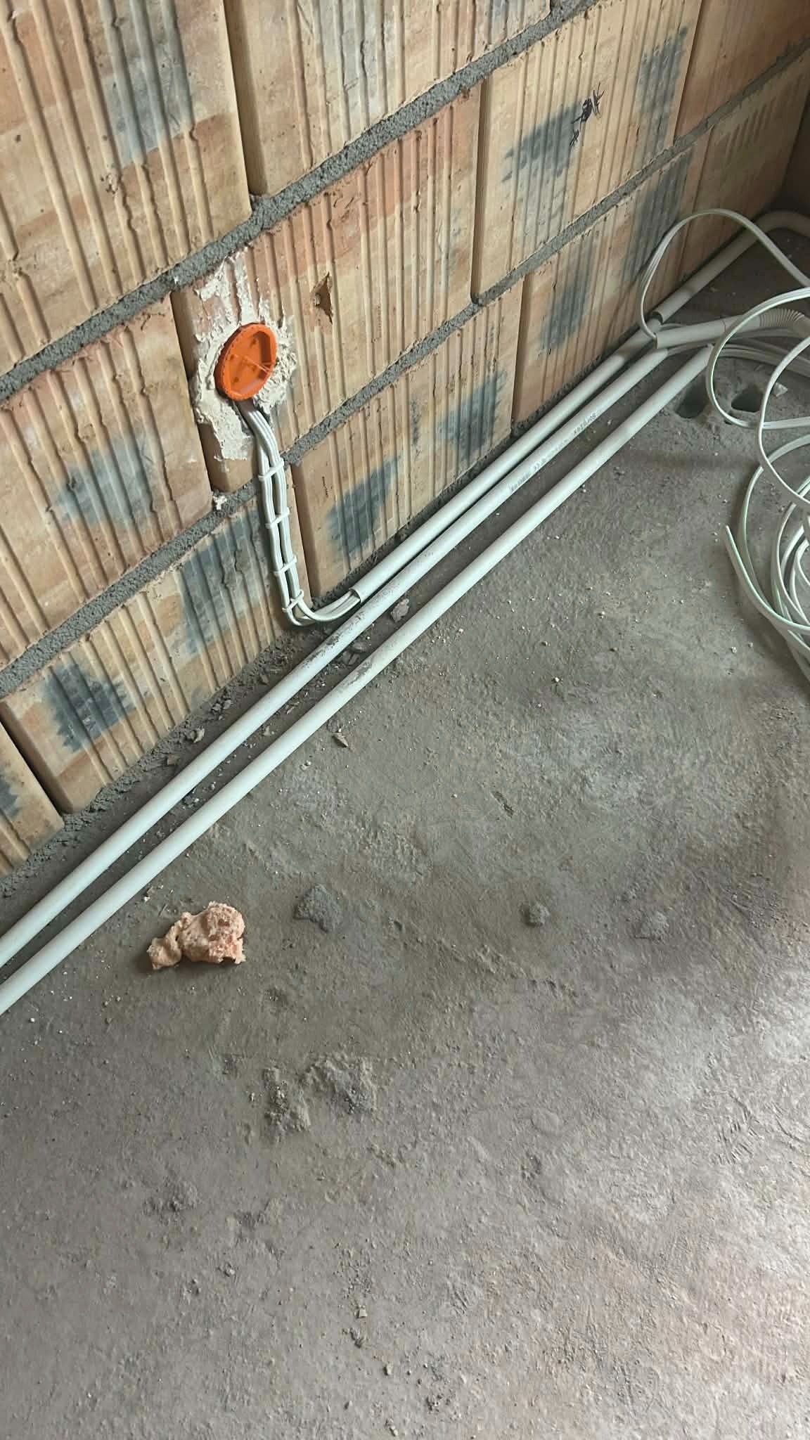 Pomarańczowa puszka instalacyjna w surowej ceglanej ścianie z białymi przewodami i rurami PCV na szarym betonowym podłożu. Widok z góry, etap prac instalacyjnych.
