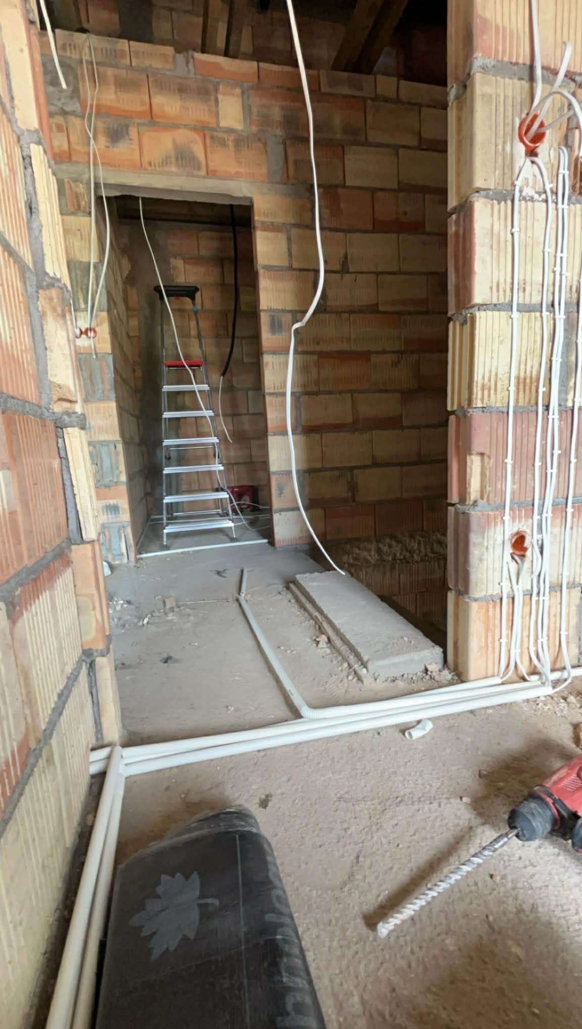 Instalacja elektryczna w surowym budynku: przewody w ścianach z pustaków, drabina w przejściu, wiertarka na podłodze. Widok na wczesny etap prac.