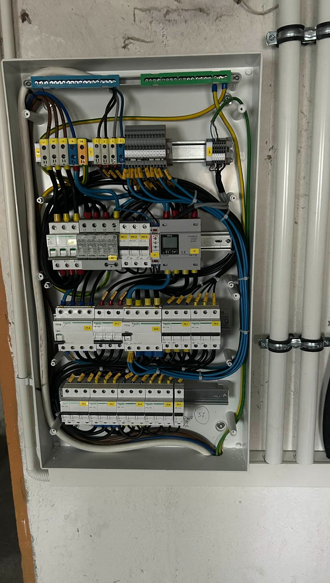 Rozdzielnica elektryczna w białej obudowie, widoczne liczne przewody, bezpieczniki i moduły. Instalacja wewnątrz skrzynki, z detalami okablowania i oznaczeń.