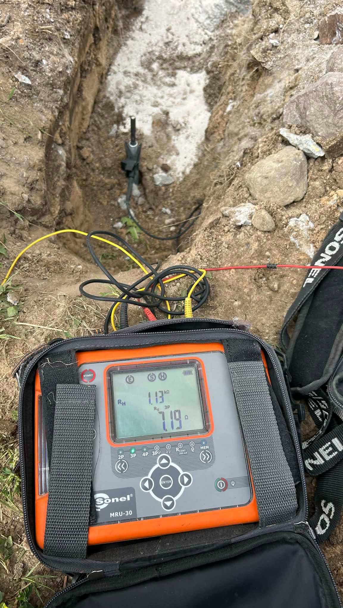 Pomiar rezystancji uziomu w wykopie za pomocą miernika Sonel MRU-30. Widoczne przewody pomiarowe i elektroda wbita w ziemię. Pomiar w trakcie realizacji.