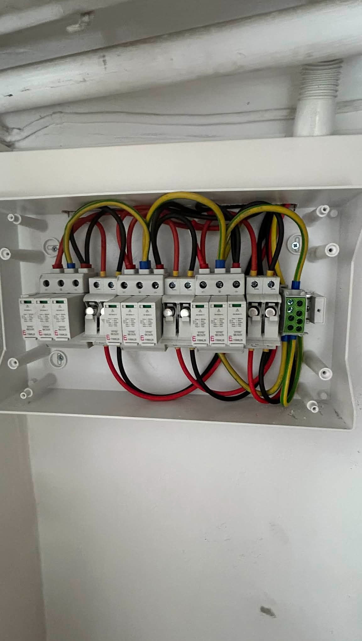 Zamknięta skrzynka z zamontowanymi bezpiecznikami i podłączonymi przewodami w kolorach: czarnym, czerwonym, żółto-zielonym. Widok z góry na białą skrzynkę.