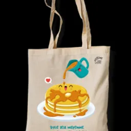 Beżowa torba bawełniana z nadrukiem graficznym: uśmiechnięte naleśniki polewane syropem z wesołego dzbanka, obok logo 'Torba Smaku' i napis 'Życie jest niepewne. Najpierw zjedz deser!'