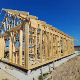 Surowy szkielet drewnianej konstrukcji domu na betonowej podstawie, widok z zewnątrz w słoneczny dzień, z widoczną konstrukcją dachu i ścian.