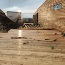 Drewniany taras w trakcie budowy, widoczne deski, narzędzia, budynek z drewnianą elewacją i konstrukcja szkieletowa w tle.