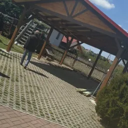 Drewniana konstrukcja z czerwonym dachem w ogrodzie, mężczyzna oddala się, widoczna drabina oparta o konstrukcję, kostka ażurowa na podłożu.