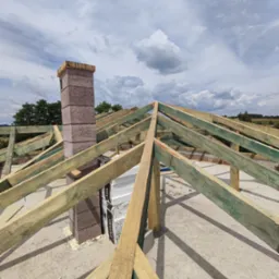 Konstrukcja drewniana dachu w trakcie budowy, widoczny komin z bloczków betonowych, niebo z chmurami w tle.