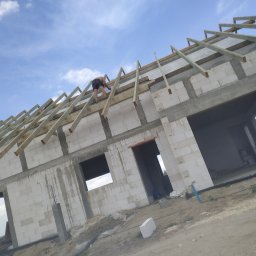 TK Ciesielstwo - Budowa domu w trakcie: murowane ściany z betonu komórkowego i montaż drewnianej konstrukcji dachu przez pracownika na tle błękitnego nieba.