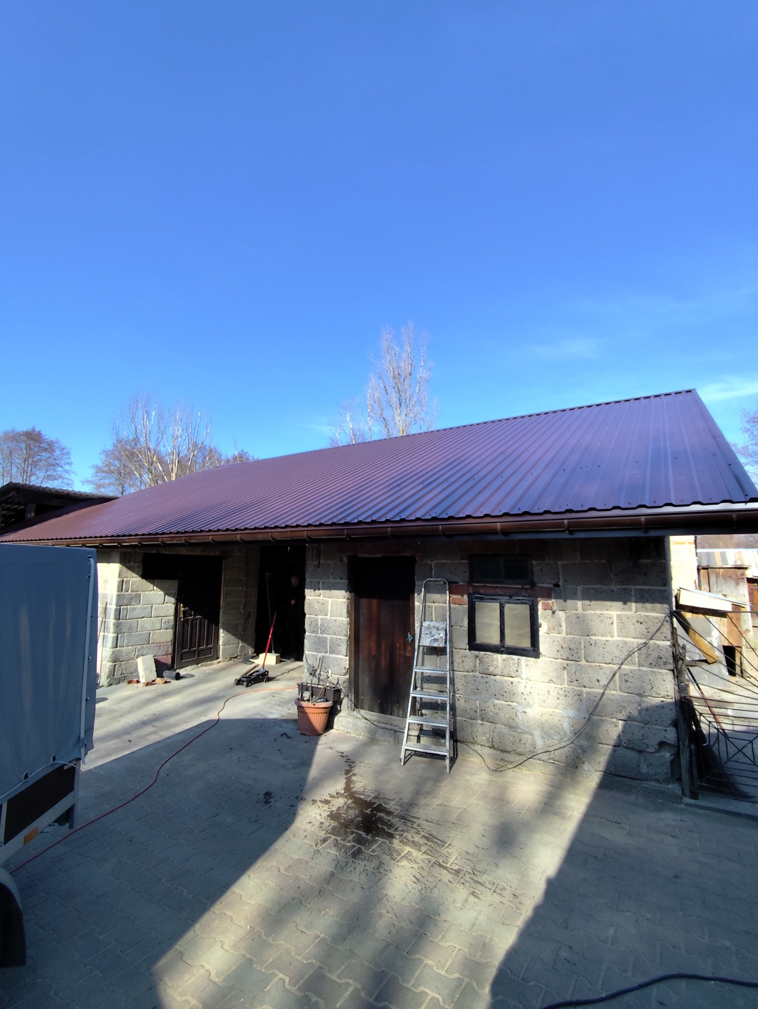 Budynek gospodarczy z nowym, metalowym dachem w kolorze bordowym, drabina oparta o ścianę, ziemia wokół budynku częściowo zabrudzona, błękitne niebo w tle.