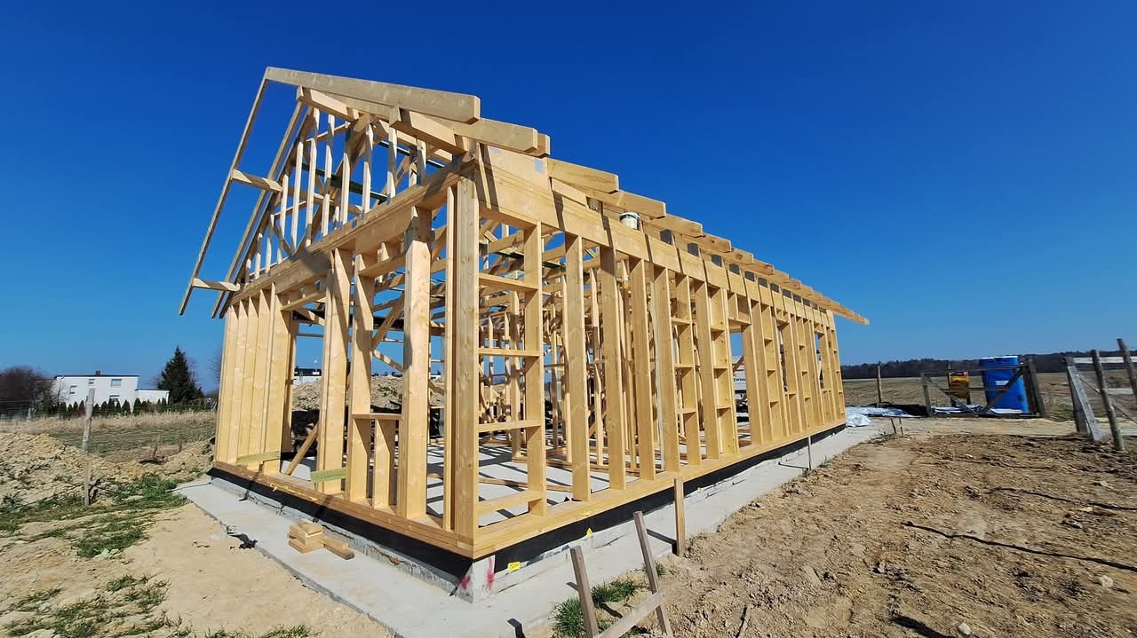 Surowy szkielet drewnianej konstrukcji domu na betonowej podstawie, widok z zewnątrz w słoneczny dzień, z widoczną konstrukcją dachu i ścian.