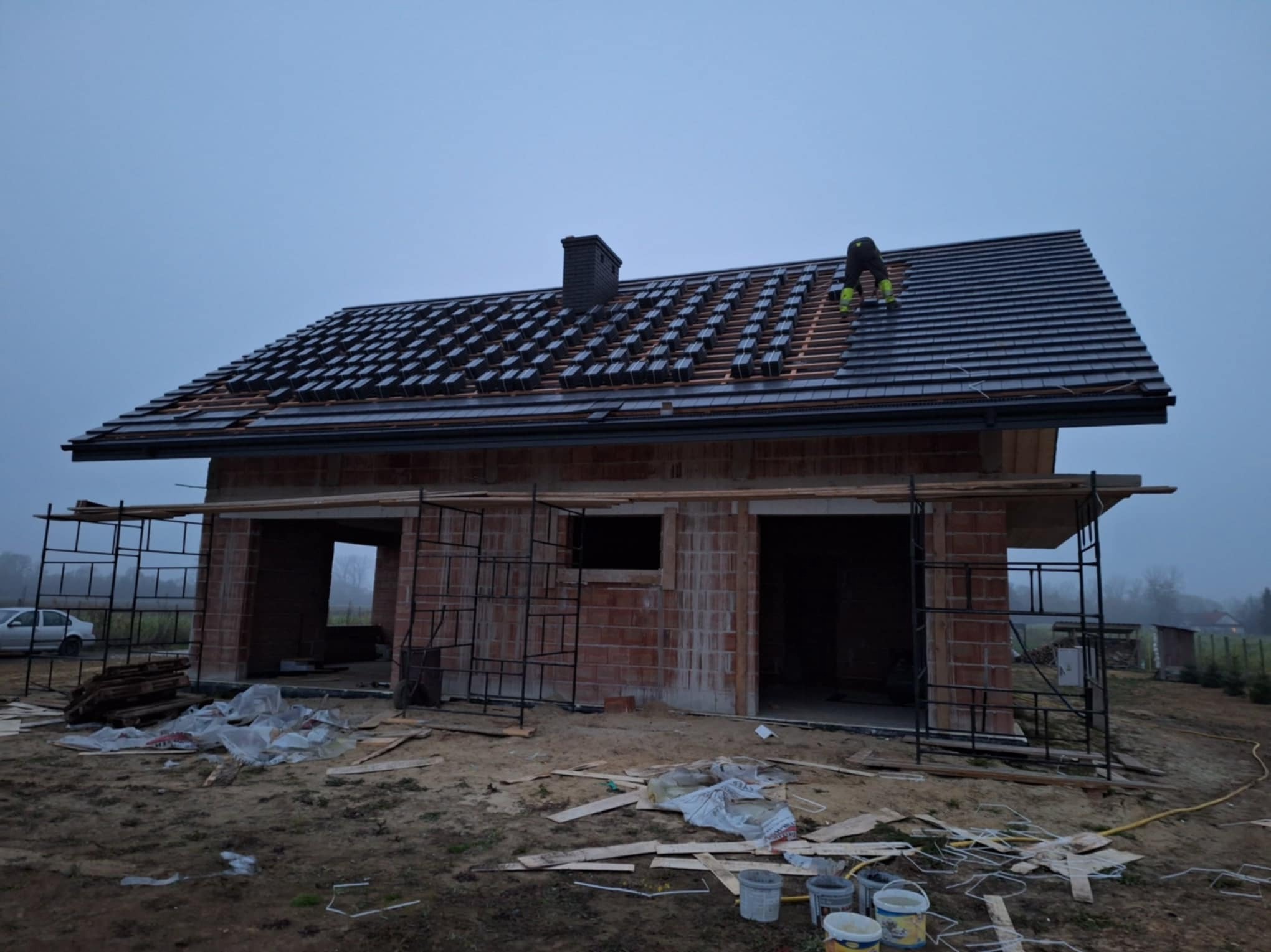 Murarz na dachu krytym częściowo czarną dachówką, widoczne rusztowania i niedokończone mury budynku w pochmurny dzień.