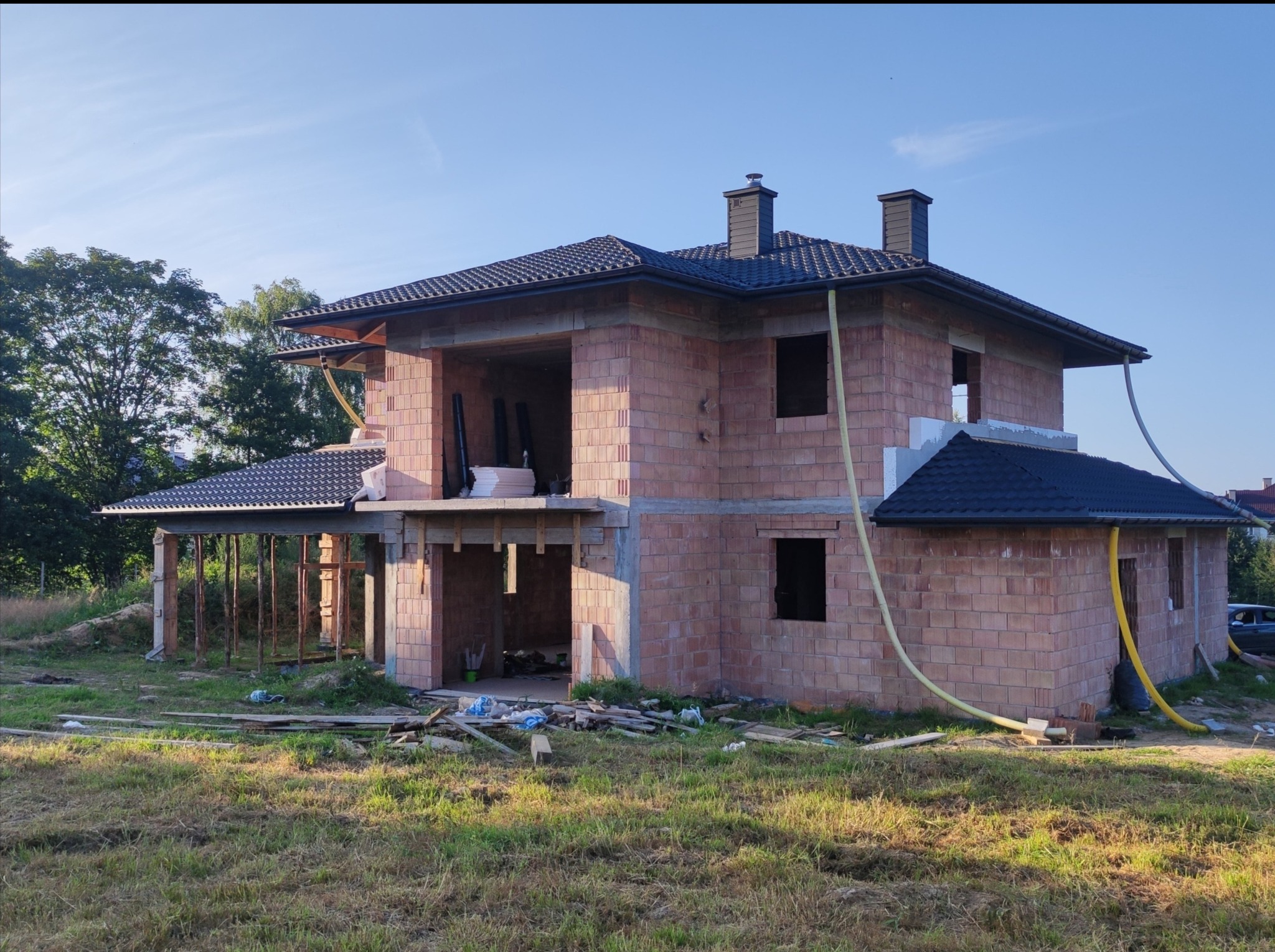 Niedokończony dom murowany z cegły ceramicznej z ciemną dachówką, widoczne rusztowania drewniane pod zadaszeniem tarasu, przewody wentylacyjne i fragmenty materiałów budowlanych na trawie.