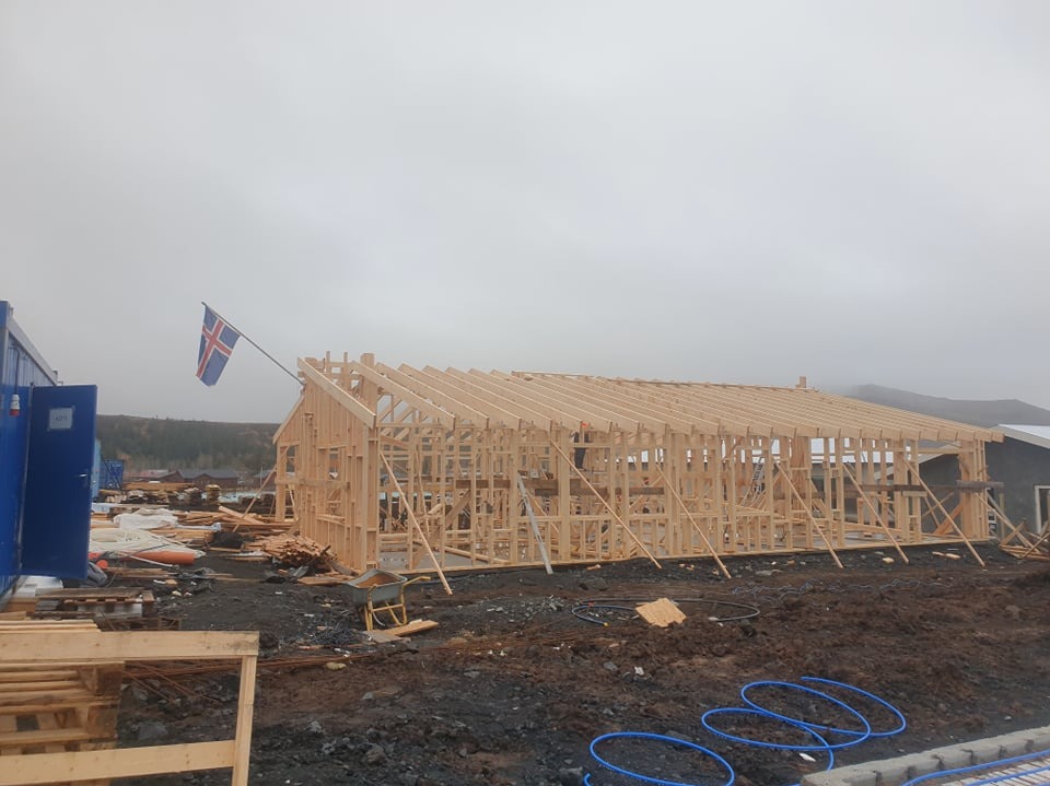 Wznoszenie drewnianej konstrukcji domu w systemie szkieletowym, na placu budowy powiewa flaga Islandii, widoczne materiały budowlane i narzędzia.