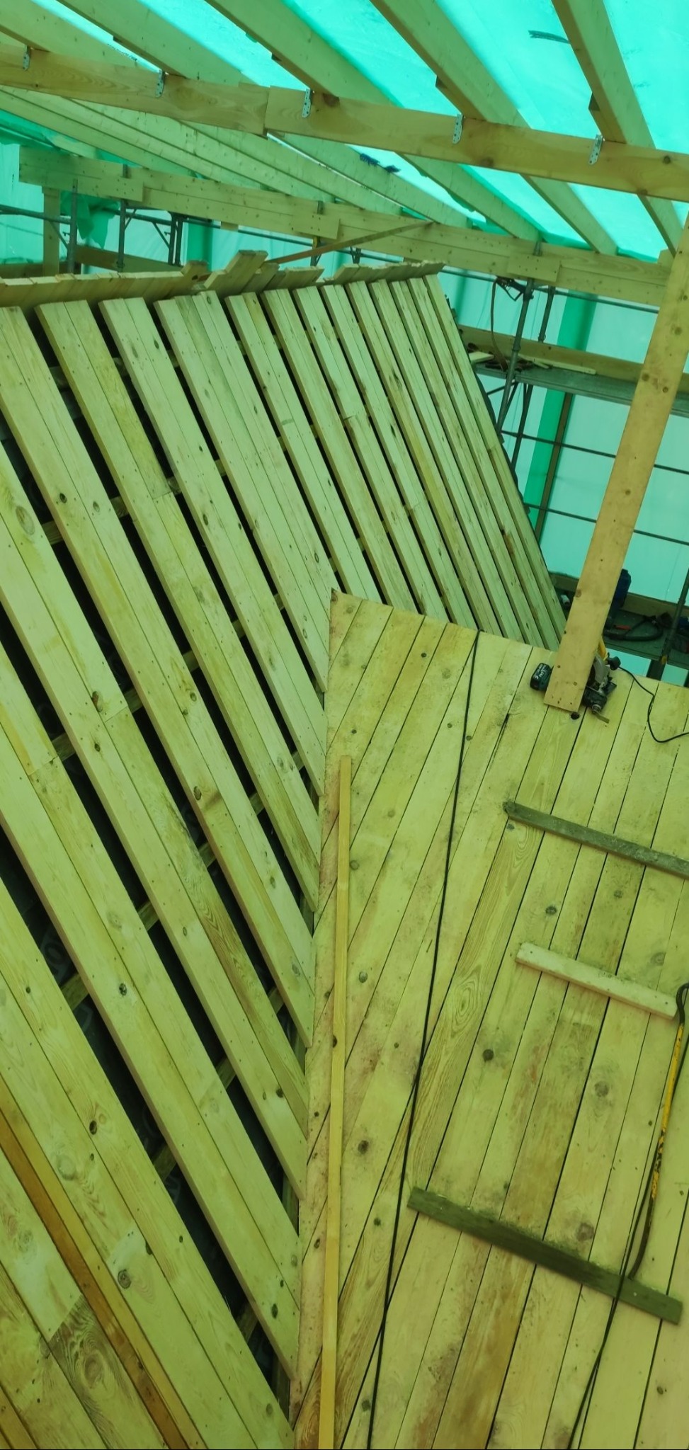 Wewnętrzny widok na konstrukcję dachu w trakcie budowy, z widocznymi elementami drewnianymi, deskami ułożonymi pod kątem i narzędziami leżącymi na powierzchni, całość przykryta zieloną plandeką.