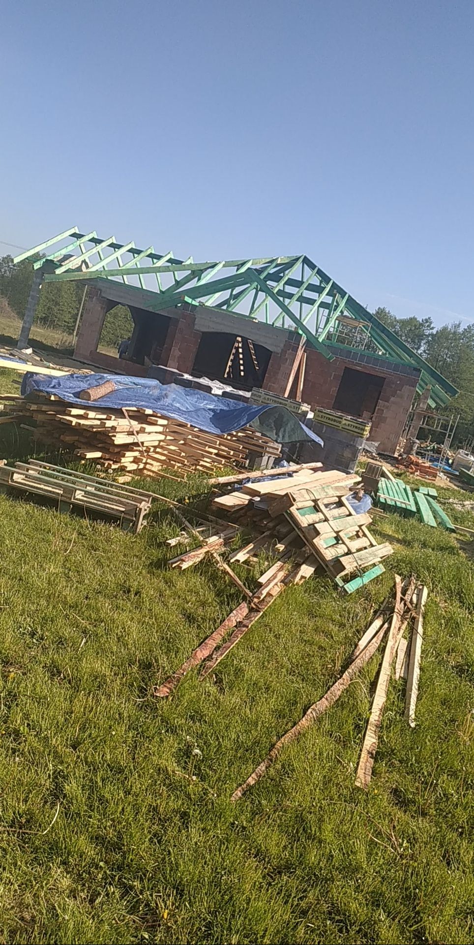 Wznoszenie więźby dachowej z zielonych belek na murowanym budynku w trakcie budowy, z widocznymi stertami drewna i rusztowaniami w tle.