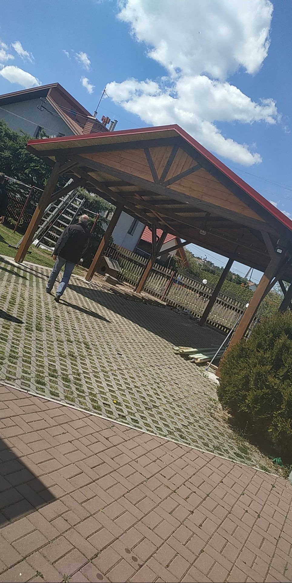 Drewniana konstrukcja z czerwonym dachem w ogrodzie, mężczyzna oddala się, widoczna drabina oparta o konstrukcję, kostka ażurowa na podłożu.