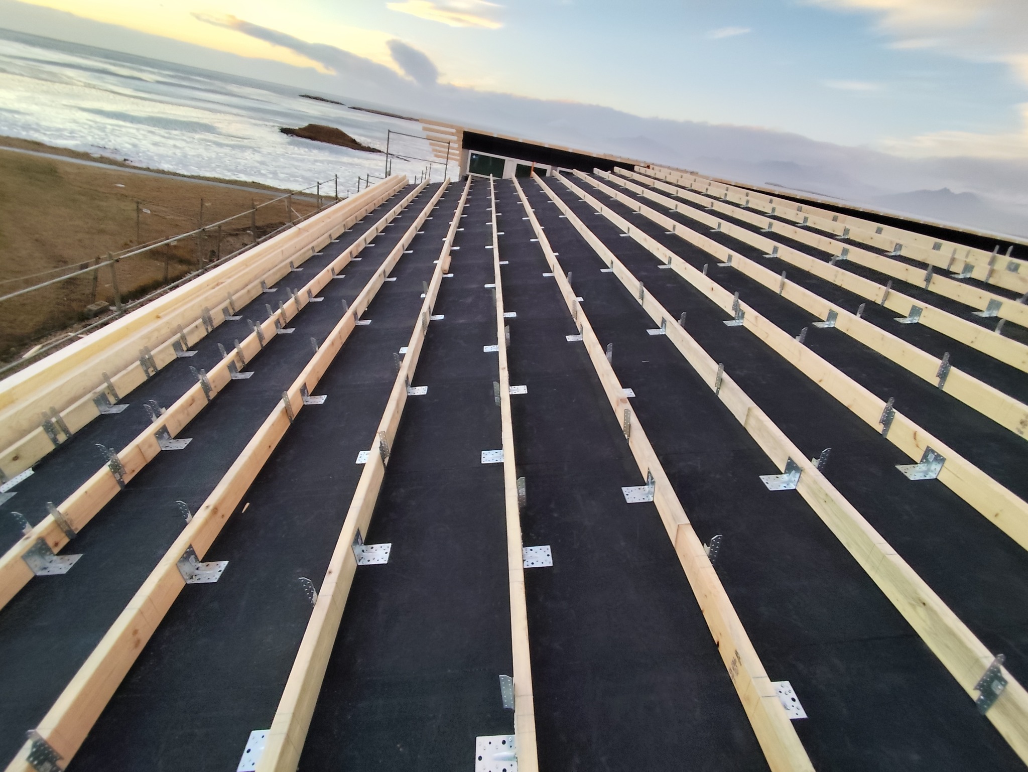 Wstępny etap budowy dachu z widoczną więźbą dachową, czarną membraną i metalowymi łącznikami, w tle krajobraz z wodą i zachodzącym słońcem.