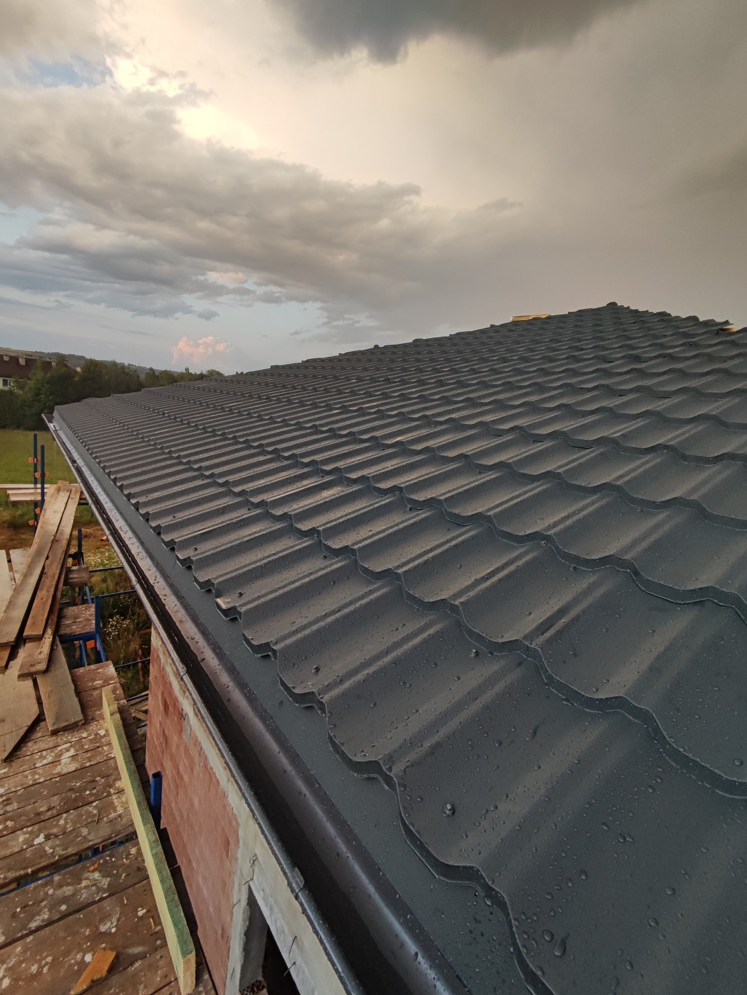 Nowy, ciemnoszary dach z blachy trapezowej, pokryty kroplami deszczu, widoczny z rusztowania, z fragmentem muru i rynny, na tle pochmurnego nieba.