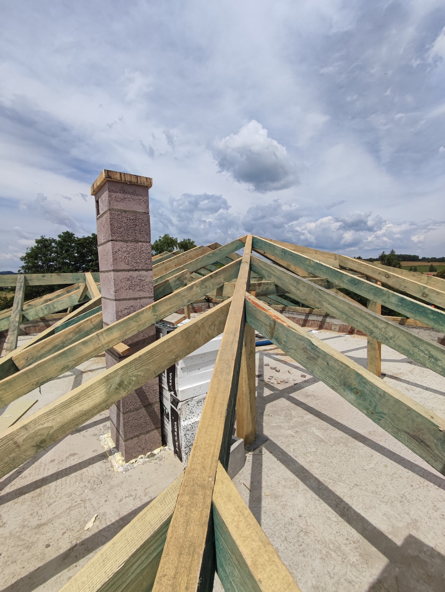 Konstrukcja drewniana dachu w trakcie budowy, widoczny komin z bloczków betonowych, niebo z chmurami w tle.