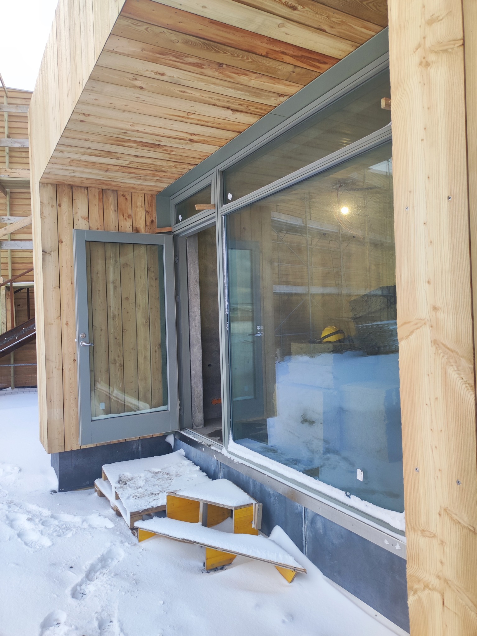 Surowy stan domu szkieletowego z drewnianą elewacją, widoczne otwarte drzwi i duże okno od zewnątrz, pokryte śniegiem prowizoryczne schody wejściowe, w tle rusztowanie budowlane.