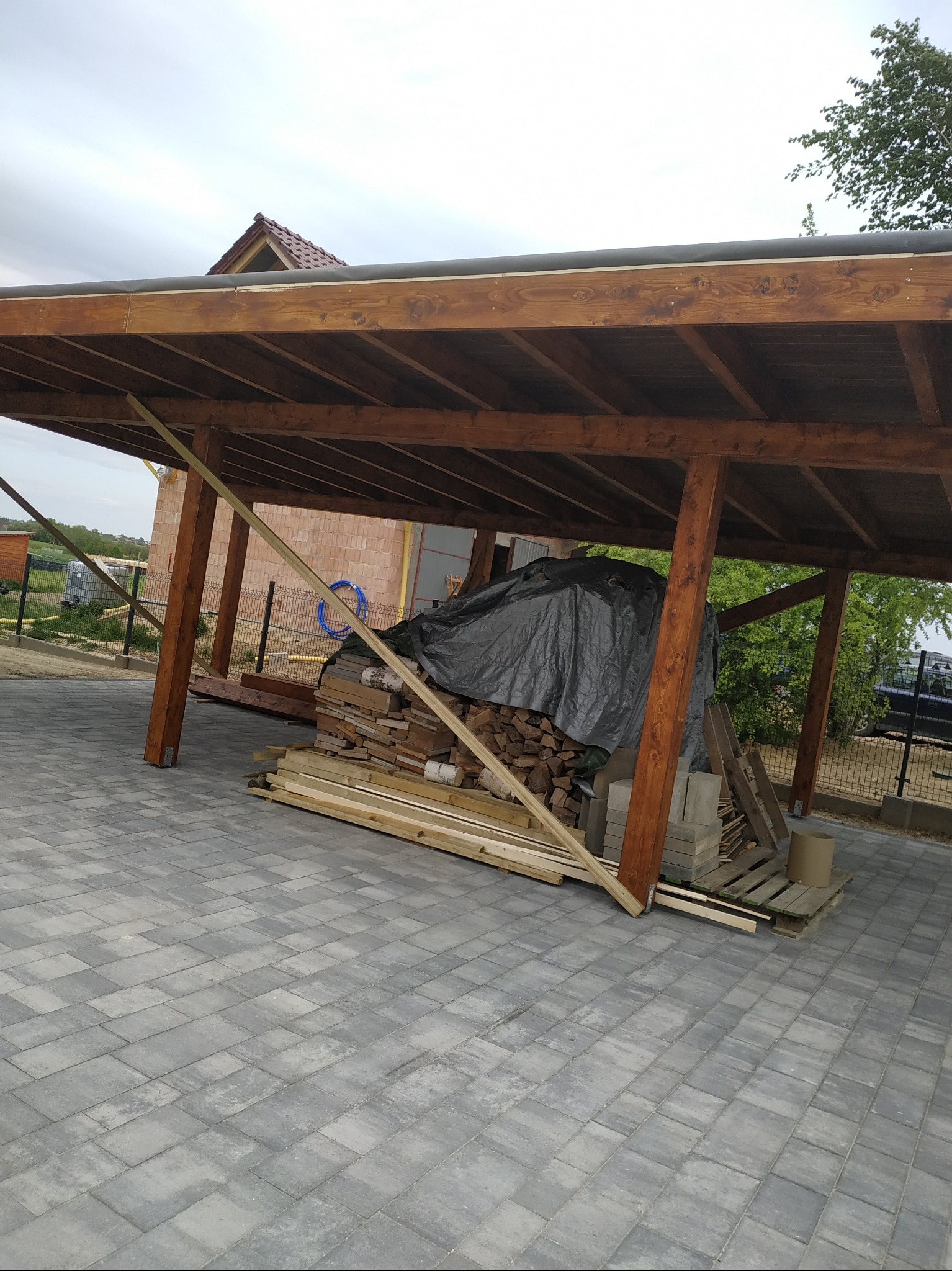 Drewniana wiata z widoczną konstrukcją dachu, pod którą składowane jest drewno opałowe przykryte szarą plandeką, obok leżą deski i kostka brukowa.