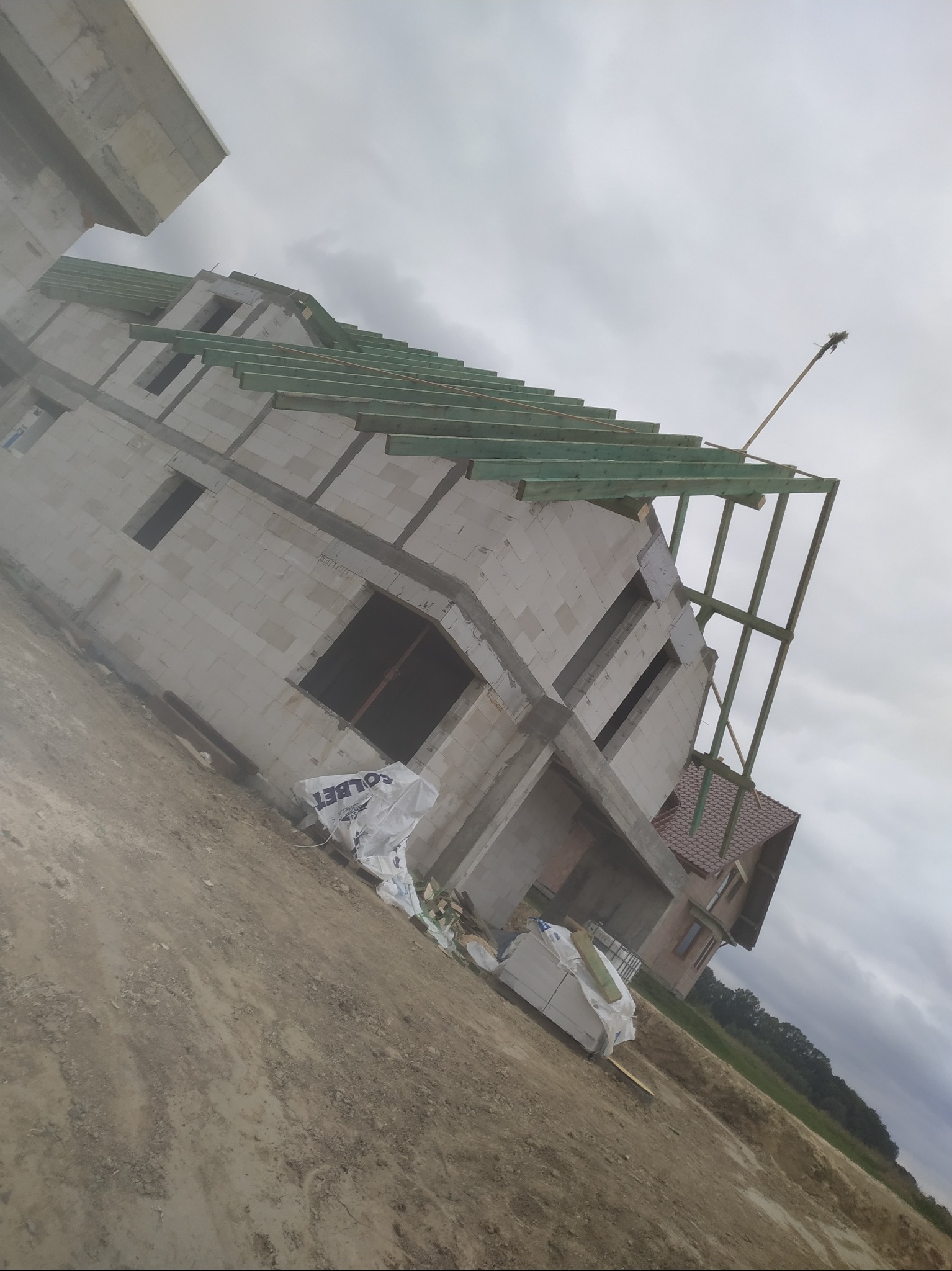 Wznoszenie domu z bloczków betonowych: widoczna konstrukcja ścian, więźba dachowa w trakcie montażu, materiały budowlane Solbet na placu budowy, sąsiedni dom z czerwonym dachem w tle.