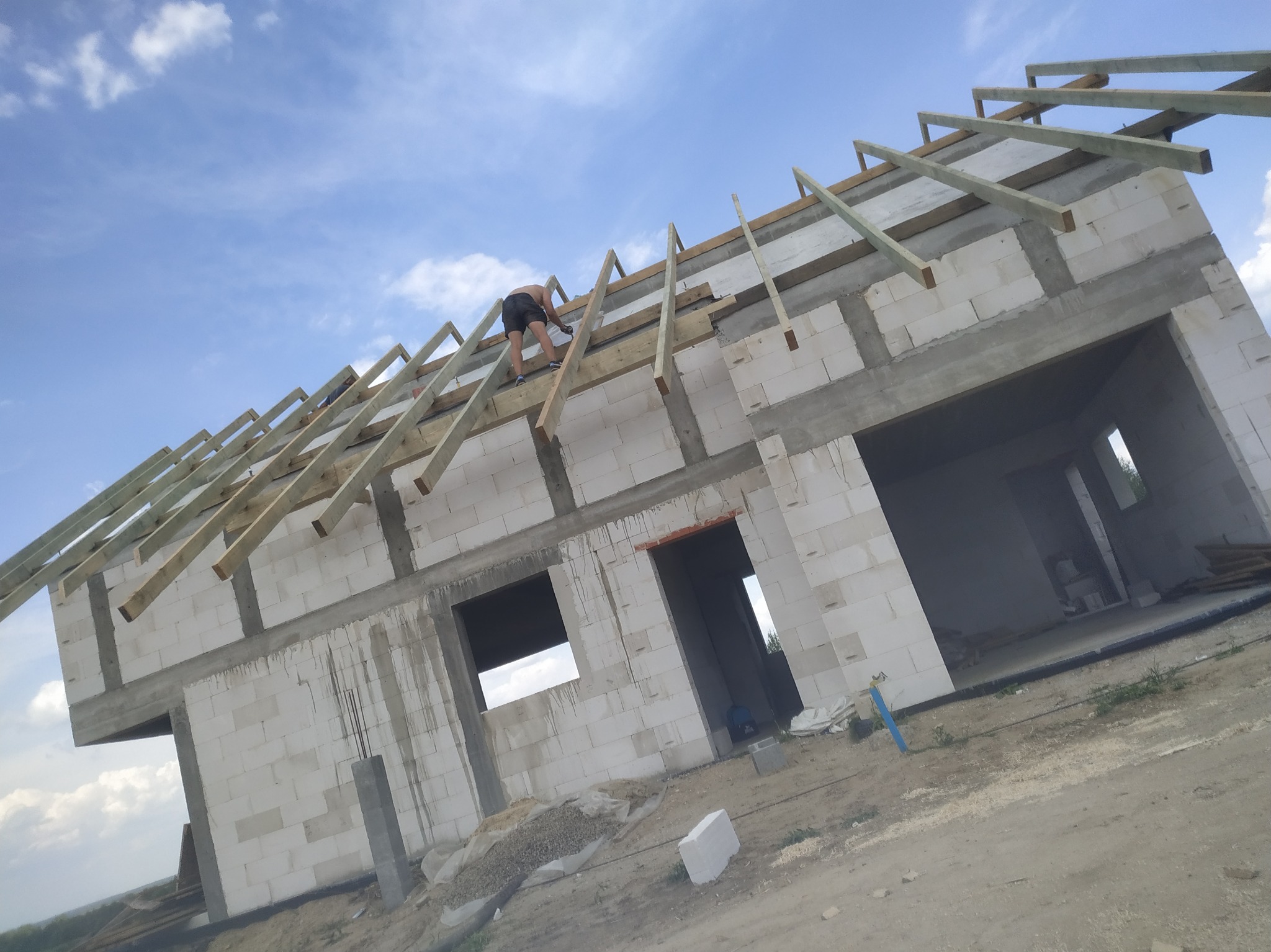 Budowa domu w trakcie: murowane ściany z betonu komórkowego i montaż drewnianej konstrukcji dachu przez pracownika na tle błękitnego nieba.