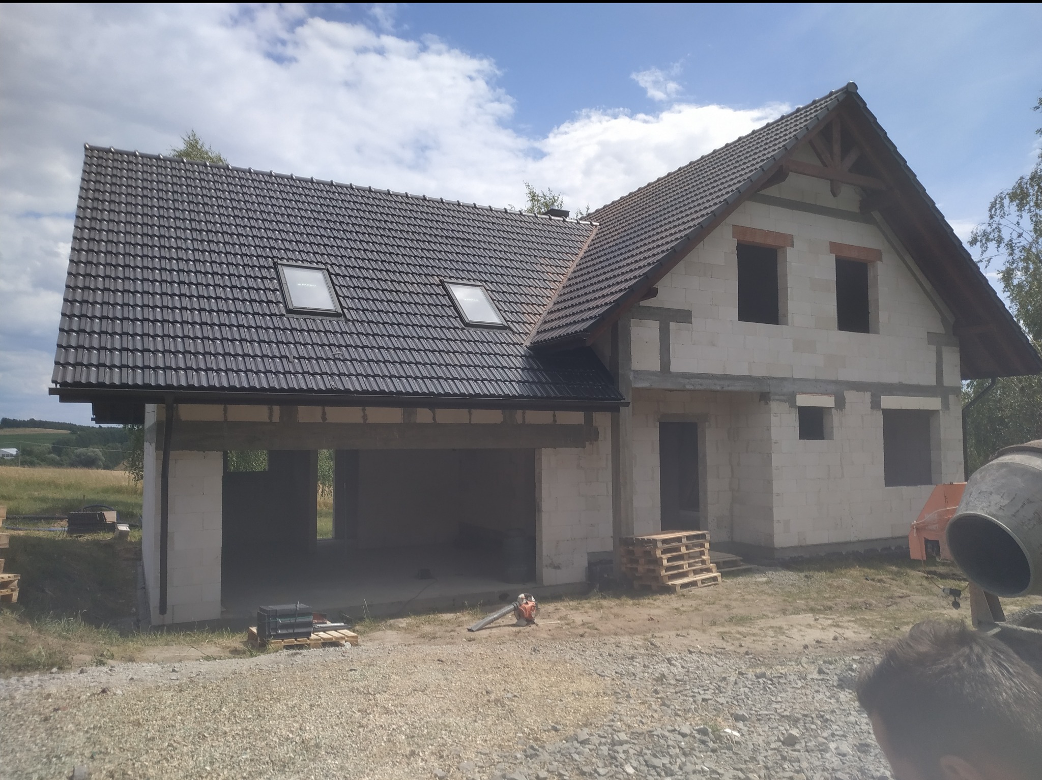 Budowa domu w trakcie realizacji, widoczna konstrukcja z białych bloczków, ciemna dachówka z oknami dachowymi, elementy drewniane więźby dachowej, teren budowy z paletami i betoniarką.