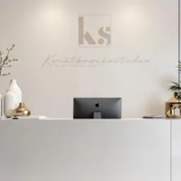 Recepcja biura z logo 'KS' i napisem 'Kwiatkowska Studio Projektowanie Wnętrz' na ścianie, minimalistyczny design, białe i złote akcenty dekoracyjne, monitor Apple w tle.