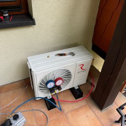 EkoKlima Montaż klimatyzacji - Biała, zewnętrzna jednostka klimatyzacji Rotenso, z podłączonymi manometrami i przewodami, umieszczona na balkonie z brązowymi płytkami, z widocznym kluczem nastawnym na obudowie i narzędziami...