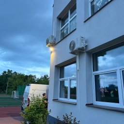 EkoKlima Montaż klimatyzacji - Biały budynek z dwoma zamontowanymi klimatyzatorami na ścianie zewnętrznej, widoczne ozdoby okienne dla dzieci, na tle zachmurzone niebo i zaparkowany biały samochód dostawczy.