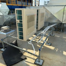 EkoKlima Montaż klimatyzacji - Dwa urządzenia klimatyzacyjne marki Fujitsu zamontowane na metalowej konstrukcji na dachu budynku. Widoczne białe rury i przewody, elementy wentylacyjne w tle.
