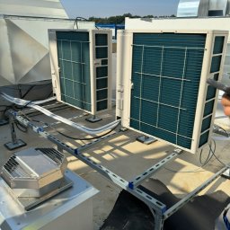 EkoKlima Montaż klimatyzacji - Dwa klimatyzatory przemysłowe zamontowane na dachu budynku, połączone systemem rur i metalowych wsporników. Widoczny fragment dachu z elementami wentylacji.