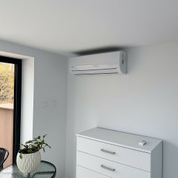 EkoKlima Montaż klimatyzacji - Biała klimatyzacja ścienna zamontowana w minimalistycznym, jasnym wnętrzu domu z białymi ścianami, komodą i rośliną w ozdobnej doniczce na szklanym stoliku.