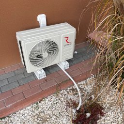 EkoKlima Montaż klimatyzacji - Zewnętrzna jednostka klimatyzacji Rotenso zamontowana na brązowej ścianie domu, widoczna z boku, z przewodami i elementami montażowymi na tle kamieni ozdobnych i roślinności.