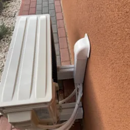 Zewnętrzna jednostka klimatyzatora zamontowana na ścianie budynku z pomarańczową elewacją, widoczne kable w plastikowej osłonie, obok kostka brukowa i kamyki.