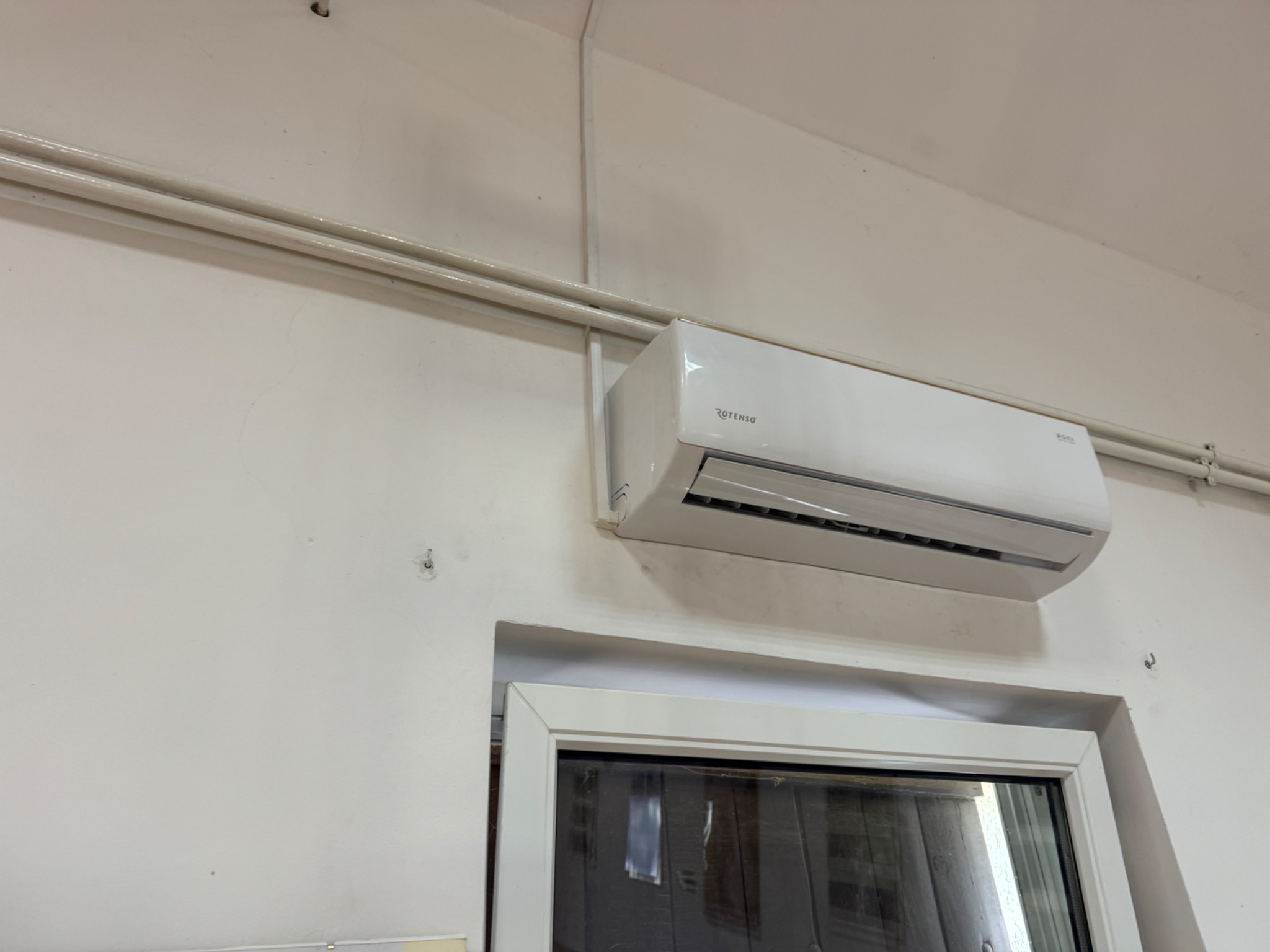 Biały klimatyzator marki Rotenso zamontowany na ścianie nad oknem, widoczne rury instalacyjne.