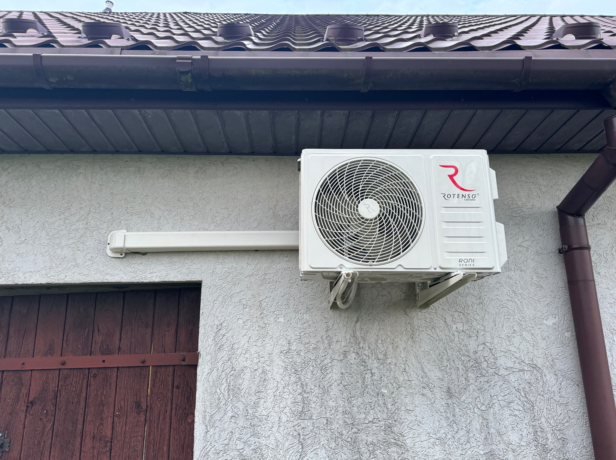 Biała jednostka zewnętrzna klimatyzatora Rotenso Roni Series zamontowana na elewacji budynku z brązowymi elementami dachu i rynny.