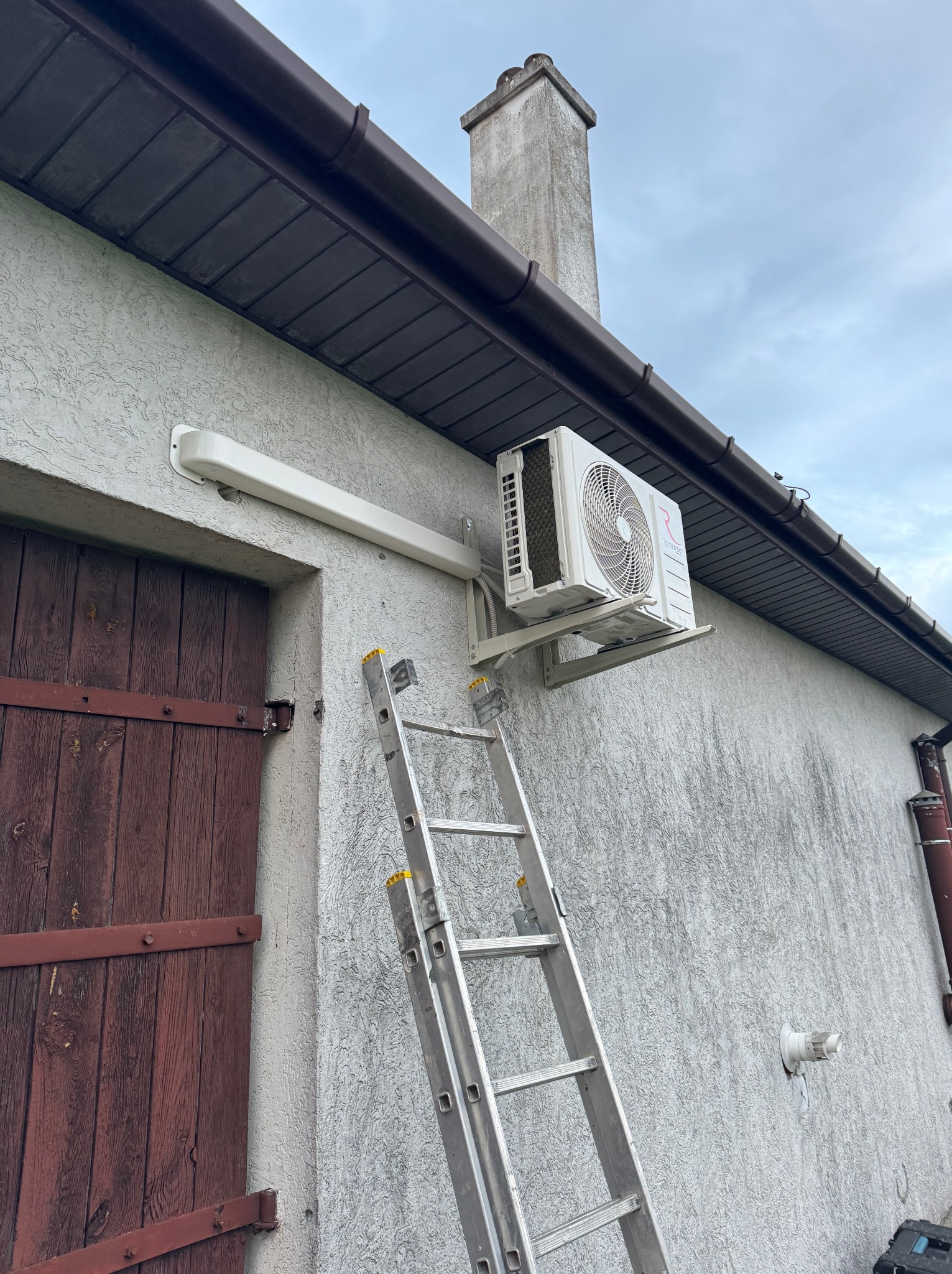 Zewnętrzna jednostka klimatyzacji zamontowana na elewacji budynku z drabiny przystawionej do ściany, widoczny komin i brązowe okiennice.
