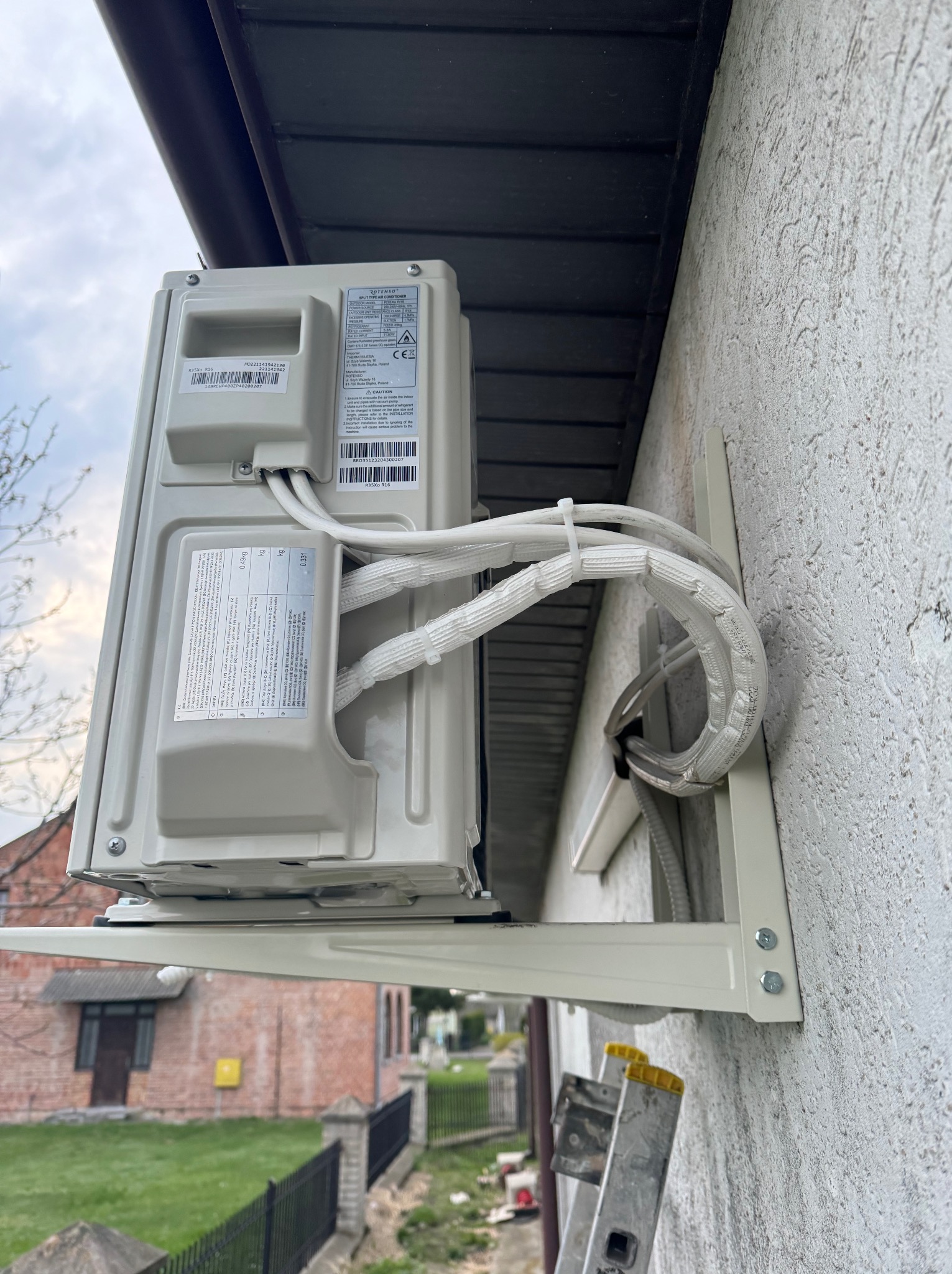 Zewnętrzna jednostka klimatyzatora zamontowana na ścianie budynku, widoczne rury w izolacji termicznej przymocowane do ściany, drabina oparta o ścianę w tle.