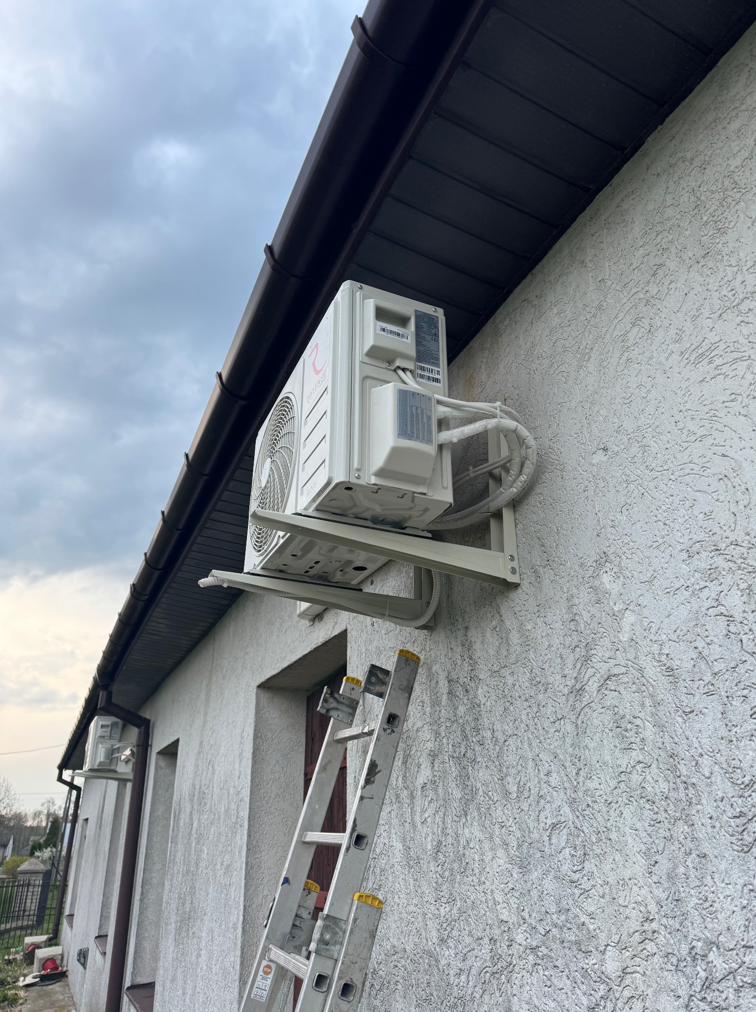 Zewnętrzna jednostka klimatyzacji zamontowana na ścianie domu z fakturą, widoczna drabina oparta o ścianę, pochmurne niebo w tle.