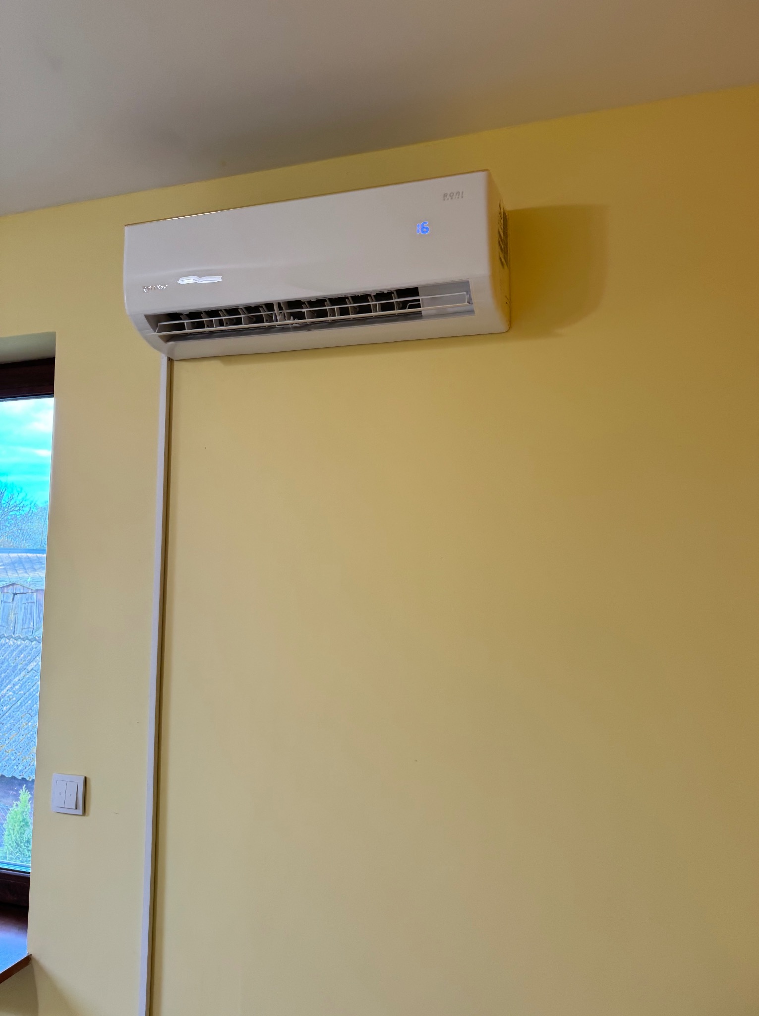 Biała jednostka klimatyzacyjna zamontowana na żółtej ścianie w pokoju, z widocznym oknem w tle.