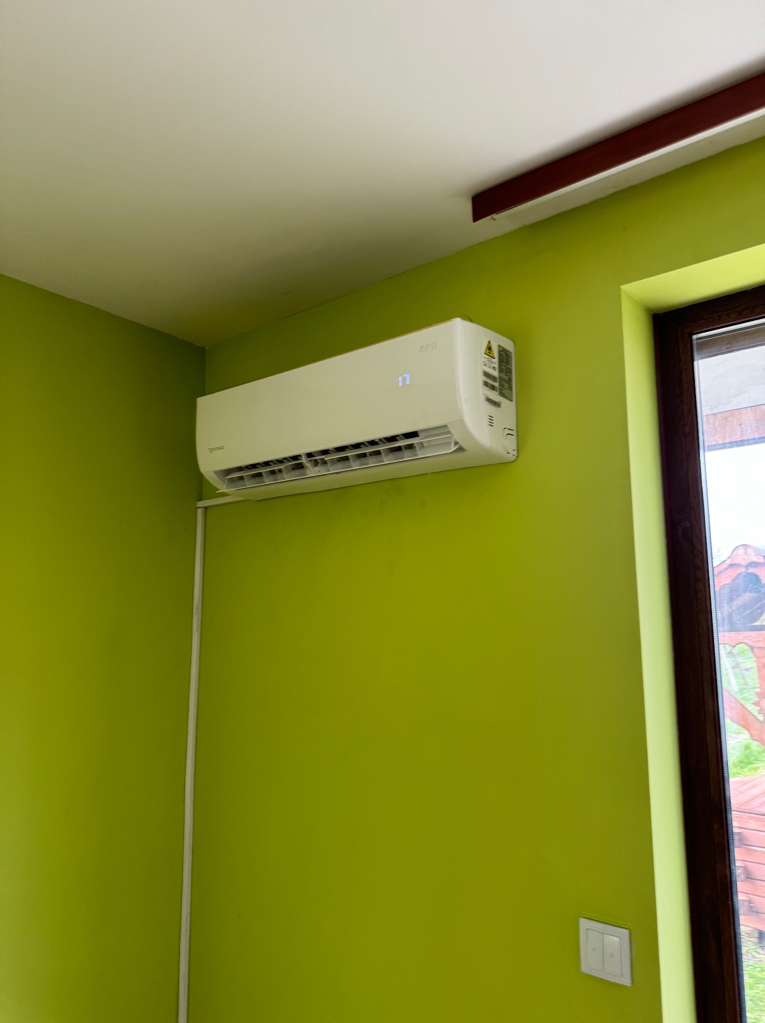 Biały klimatyzator na zielonej ścianie w pomieszczeniu, widoczny kabel zasilający i fragment okna z drewnianą ramą.