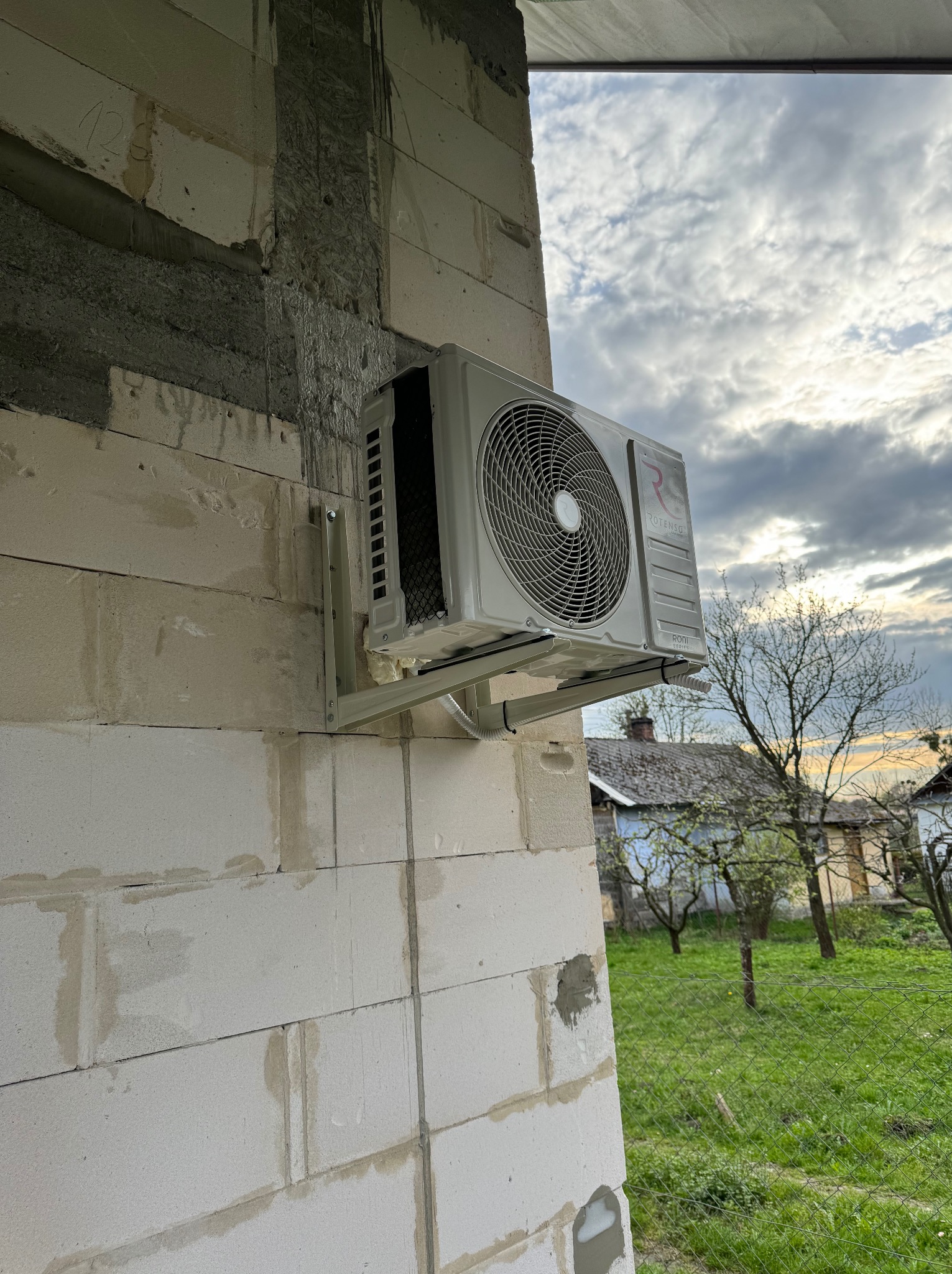 Zamontowana jednostka zewnętrzna klimatyzatora Rotenso na ścianie budynku z betonu komórkowego, widok na ogród i zachmurzone niebo.
