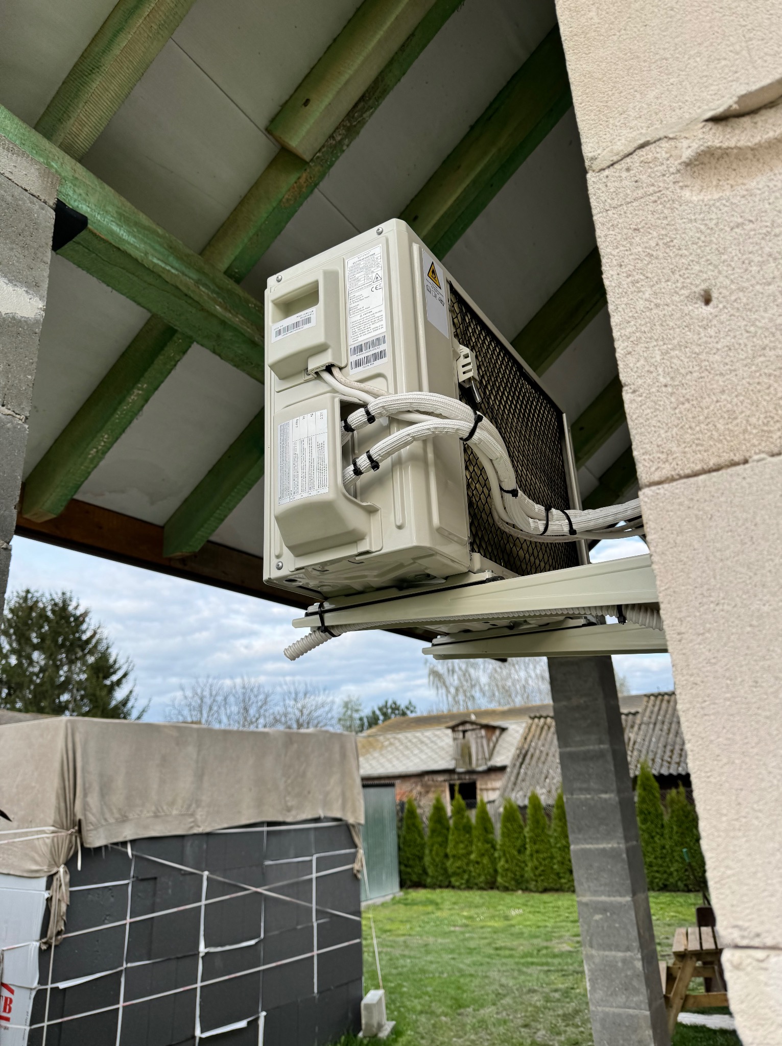 Zewnętrzna jednostka klimatyzacji zamontowana na elewacji domu w trakcie instalacji, widoczne kable w osłonach, elementy konstrukcji dachu i fragment ogrodu.